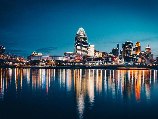A photo of downtown Cincinnati, Ohio at night. Lights from the buildings reflect on the Ohio river. 
