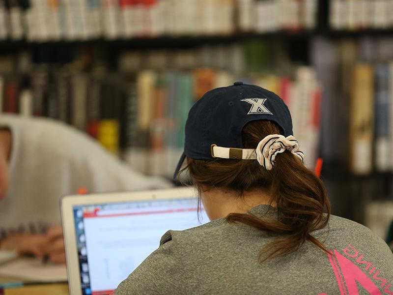 一张学生后脑勺的照片. 这个学生坐在图书馆的桌子上. 学生面前有一台开着的笔记本电脑. 背景里有几书架的书.